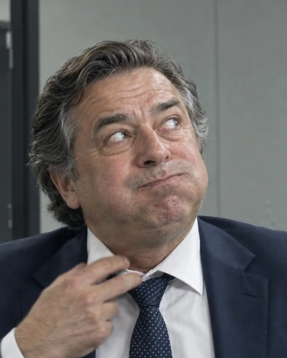 Un hombre de unos sesenta años con traje azul marino y corbata, mirando de reojo hacia arriba con la boca torcida, mientras se afloja el nudo con dos dedos.