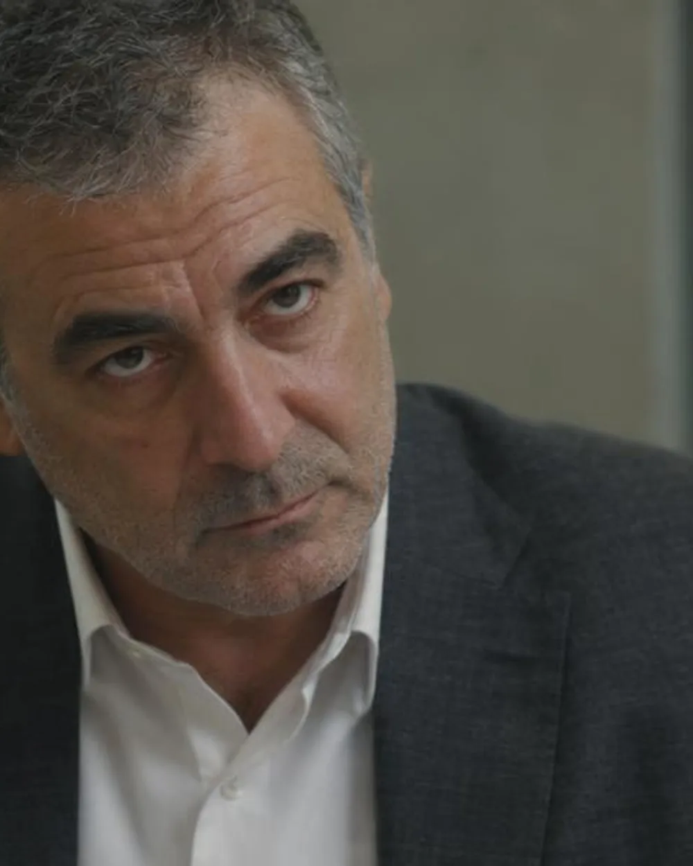 Un hombre de unos sesenta años con americana gris y camisa blanca sin corbata, la barbilla algo baja, mirando de frente por debajo de las cejas al otro lado de una mesa.