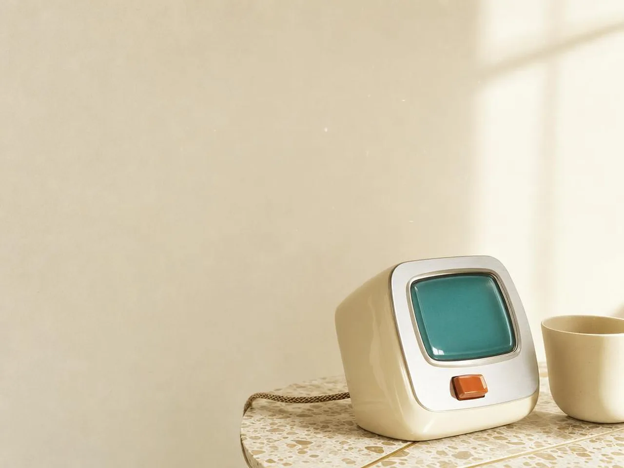 Intercomunicador de sobremesa de los años sesenta en esmalte crema, con la pantalla encendida y un pulsador naranja, sobre una mesa de terrazo junto a una taza.