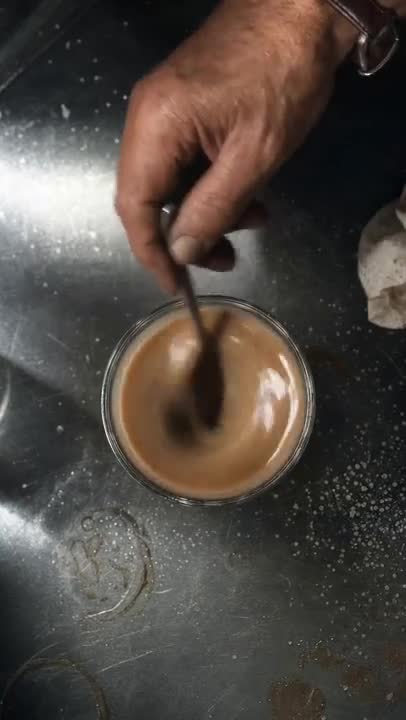 Plano cenital de un vaso de café con una mano removiendo con una cucharilla sobre una superficie metálica.